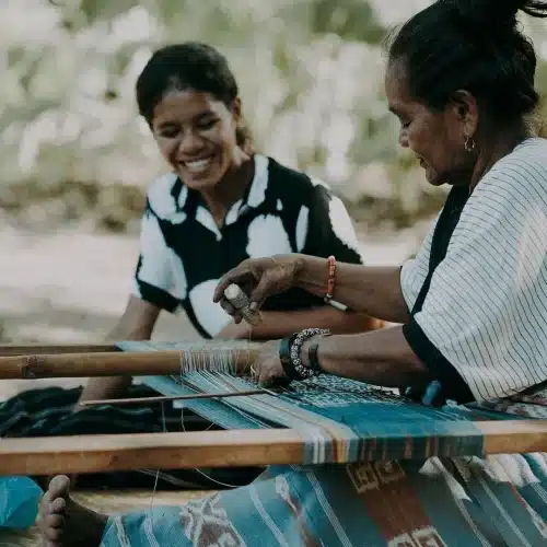 Two Sumbanese women weaving ikat cloth on a backstrap loom, symbolizing tradition and women’s empowerment.