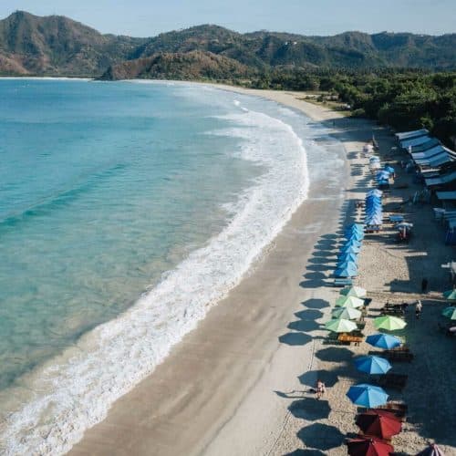 Aerial view of Selong Belanak Beach in Lombok with turquoise water, gentle waves and a line of colorful umbrellas and beach huts.