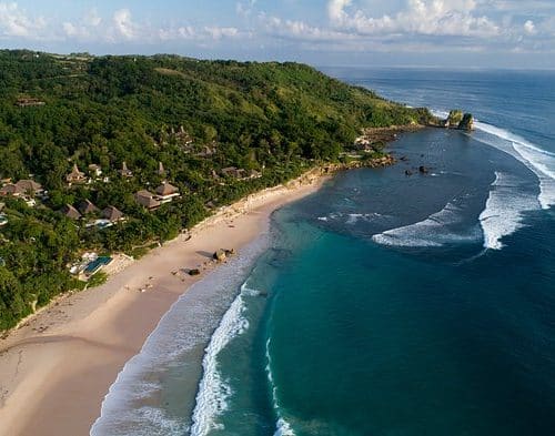 Aerial view of Nihi Sumba resort hidden in lush hills above a long empty beach on Sumba, Indonesia.